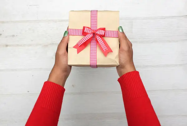 view from above ofgirl asian hands in red sweater holding craft paper gift box with red ribbon bow on a white wooden table.for giving in special day,Christmas, new year,Valentine