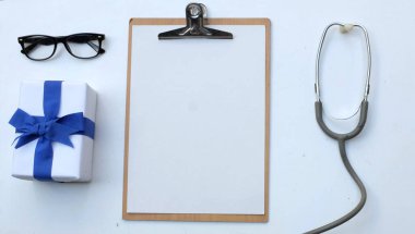 Üst ve düz, uzay doktorlarının ofis masasının fotokopisi. Tıbbi aksesuarlar, boş tıbbi pano, steteskop ve mavi kurdeleli el işi kağıt kutu, gözlük, beyaz perde arkasında kalem izolasyonu.