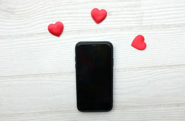A black smartphone,phone ,mobile with a heart icon, surrounded by small red hearts isolate on a wooden table white backdrop .minimalist trend.valentines day,dating mobile application, digital online
