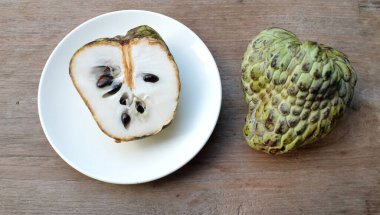 Taze yeşil kremalı elma, elma şekeri, annona cherimola (annona squamosa l..), tahta masa arkasında beyaz bir tabakta yapraklarla ikiye bölünmüş meyve. Tropikal egzotik meyve ve sağlık