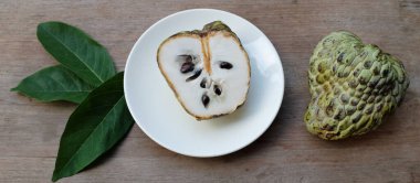 Taze yeşil kremalı elma, elma şekeri, annona cherimola (annona squamosa l..), tahta masa arkasında beyaz bir tabakta yapraklarla ikiye bölünmüş meyve. Tropikal egzotik meyve ve sağlık