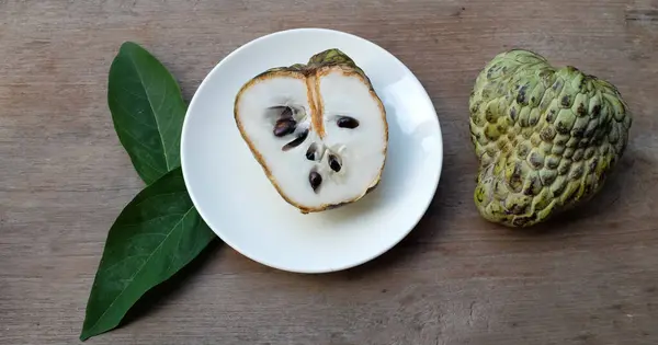 stock image fresh green raw custard apple,sugar apple, annona cherimola (annona squamosa l.) fruit cut in half sliced with leaves in a white plate on wooden table backdrop. tropical exotic fruit and health