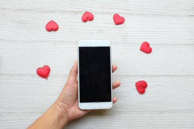 Akıllı telefon tutan Asyalı bir kadın ve beyaz ahşap bir arka planda izole edilmiş bir sürü kırmızı kalp. Teknoloji konsepti iletişim ve iş dünyası, Sevgililer Günü