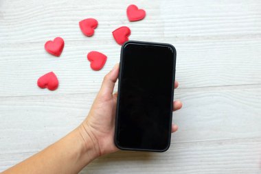 Akıllı telefon tutan Asyalı bir kadın ve beyaz ahşap bir arka planda izole edilmiş bir sürü kırmızı kalp. Teknoloji konsepti iletişim ve iş dünyası, Sevgililer Günü