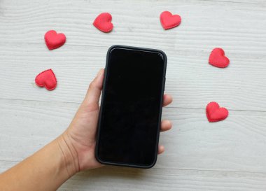 Akıllı telefon tutan Asyalı bir kadın ve beyaz ahşap bir arka planda izole edilmiş bir sürü kırmızı kalp. Teknoloji konsepti iletişim ve iş dünyası, Sevgililer Günü
