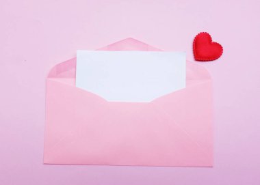 İçinde boş beyaz bir not olan pembe kağıt zarf ve dekoratif kırmızı kalp şekli, pembe pastel arka planda kalem kırmızısı. Mesajın için. Aşk mesajı, Romantik aşk, ofis kırtasiyesi ve Sevgililer Günü 