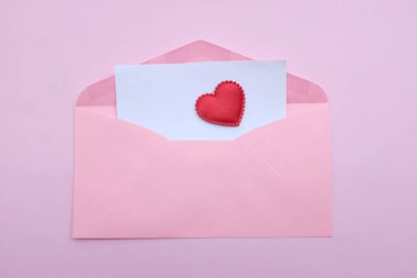 İçinde boş beyaz bir not olan pembe kağıt zarf ve dekoratif kırmızı kalp şekli, pembe pastel arka planda kalem kırmızısı. Mesajın için. Aşk mesajı, Romantik aşk, ofis kırtasiyesi ve Sevgililer Günü 