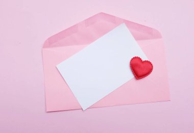 İçinde boş beyaz bir not olan pembe kağıt zarf ve dekoratif kırmızı kalp şekli, pembe pastel arka planda kalem kırmızısı. Mesajın için. Aşk mesajı, Romantik aşk, ofis kırtasiyesi ve Sevgililer Günü 