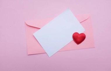 İçinde boş beyaz bir not olan pembe kağıt zarf ve dekoratif kırmızı kalp şekli, pembe pastel arka planda kalem kırmızısı. Mesajın için. Aşk mesajı, Romantik aşk, ofis kırtasiyesi ve Sevgililer Günü 
