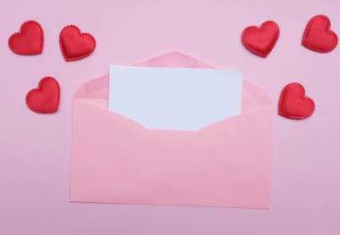İçinde boş beyaz bir not olan pembe kağıt zarf ve dekoratif kırmızı kalp şekli, pembe pastel arka planda kalem kırmızısı. Mesajın için. Aşk mesajı, Romantik aşk, ofis kırtasiyesi ve Sevgililer Günü 