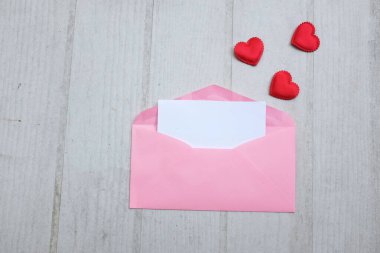 İçinde boş beyaz bir not olan pembe kağıt zarf ve dekoratif kırmızı kalp şekli, Beyaz Tahta Vintage Masa 'da kırmızı kalem. Aşk mesajı, Romantik aşk, ofis kırtasiyesi ve Sevgililer Günü 