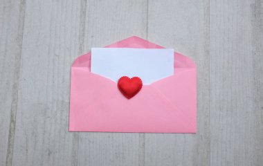 İçinde boş beyaz bir not olan pembe kağıt zarf ve dekoratif kırmızı kalp şekli, Beyaz Tahta Vintage Masa 'da kırmızı kalem. Aşk mesajı, Romantik aşk, ofis kırtasiyesi ve Sevgililer Günü 