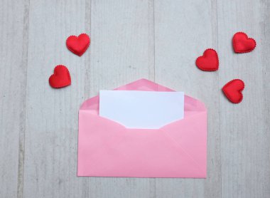 İçinde boş beyaz bir not olan pembe kağıt zarf ve dekoratif kırmızı kalp şekli, Beyaz Tahta Vintage Masa 'da kırmızı kalem. Aşk mesajı, Romantik aşk, ofis kırtasiyesi ve Sevgililer Günü 