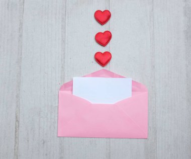 İçinde boş beyaz bir not olan pembe kağıt zarf ve dekoratif kırmızı kalp şekli, Beyaz Tahta Vintage Masa 'da kırmızı kalem. Aşk mesajı, Romantik aşk, ofis kırtasiyesi ve Sevgililer Günü 