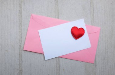 İçinde boş beyaz bir not olan pembe kağıt zarf ve dekoratif kırmızı kalp şekli, Beyaz Tahta Vintage Masa 'da kırmızı kalem. Aşk mesajı, Romantik aşk, ofis kırtasiyesi ve Sevgililer Günü 