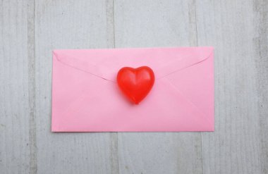 İçinde boş beyaz bir not olan pembe kağıt zarf ve dekoratif kırmızı kalp şekli, Beyaz Tahta Vintage Masa 'da kırmızı kalem. Aşk mesajı, Romantik aşk, ofis kırtasiyesi ve Sevgililer Günü 