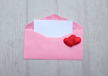İçinde boş beyaz bir not olan pembe kağıt zarf ve dekoratif kırmızı kalp şekli, Beyaz Tahta Vintage Masa 'da kırmızı kalem. Aşk mesajı, Romantik aşk, ofis kırtasiyesi ve Sevgililer Günü 