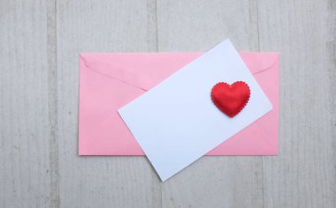 İçinde boş beyaz bir not olan pembe kağıt zarf ve dekoratif kırmızı kalp şekli, Beyaz Tahta Vintage Masa 'da kırmızı kalem. Aşk mesajı, Romantik aşk, ofis kırtasiyesi ve Sevgililer Günü 
