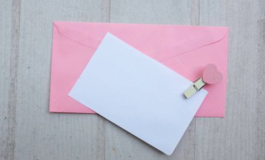 İçinde beyaz bir not olan pembe kağıt zarf. Dekoratif kalp şeklinde tahta ataç. Beyaz Ahşap Masa 'da kırmızı kalem. Aşk mesajı, Romantik aşk, ofis kırtasiyesi ve Sevgililer Günü 