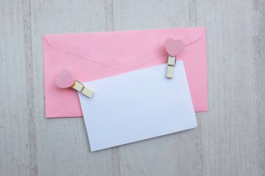 İçinde beyaz bir not olan pembe kağıt zarf. Dekoratif kalp şeklinde tahta ataç. Beyaz Ahşap Masa 'da kırmızı kalem. Aşk mesajı, Romantik aşk, ofis kırtasiyesi ve Sevgililer Günü 