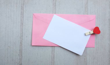 İçinde beyaz bir not olan pembe kağıt zarf. Dekoratif kalp şeklinde tahta ataç. Beyaz Ahşap Masa 'da kırmızı kalem. Aşk mesajı, Romantik aşk, ofis kırtasiyesi ve Sevgililer Günü 