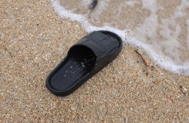 Kumlu sahilde tek bir siyah parmak arası terlik ve kıyıya vuran dalgalar. Yaz tatili ve tropikal tatil konsepti.