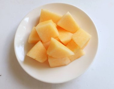 Olgun portakal sarısı yedi, sarı ballı kavun, altın karpuz parçalara ayrılmış ve beyaz seramik bir tabakta yemeye hazır beyaz bir zemin üzerinde izole edilmiş.