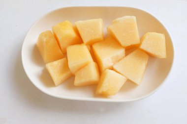 Olgun portakal sarısı yedi, sarı ballı kavun, altın karpuz parçalara ayrılmış ve beyaz seramik bir tabakta yemeye hazır beyaz bir zemin üzerinde izole edilmiş.