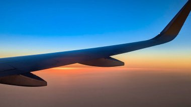 Renkli ve sembolik bir görüntü, deniz üzerinde günbatımı olan bir ticari uçağın kanadı.