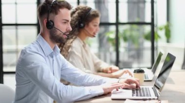 Kulaklıklı, gülümseyen erkek çağrı merkezi operatörü, modern ofiste arka planda meslektaşlarıyla oturuyor, online danışmanlık yapıyor..