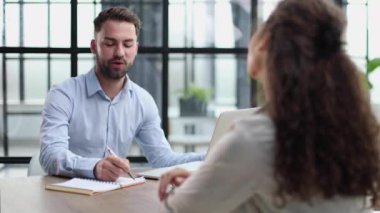 Close-up of a worker writing on a clipboard while sitting at a desk in front of a female colleague