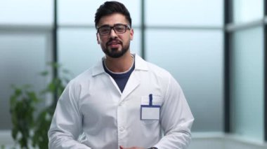Gözlüklü gülümseyen erkek doktor hastane koridorunda duruyor.