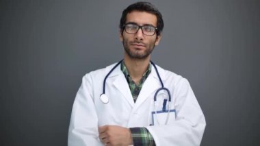 Pozitif erkek doktor portresi kollarını kavuşturup gri stüdyo arka planında kameraya gülümsüyor.