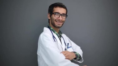 Pozitif erkek doktor portresi kollarını kavuşturup gri stüdyo arka planında kameraya gülümsüyor.