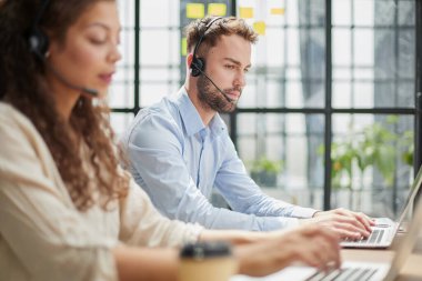 Modern ofiste kulaklıklı bir erkek çağrı merkezi operatörü, bir dizüstü bilgisayarda online bilgi danışma, müşteriye yardımcı olmak için bir dosyada bilgi arama.