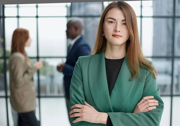 stock image businesswoman standing with her arms crossed in an office while her colleagues have a meeting in the background.