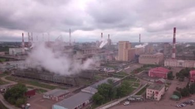 Bulutlu bir gökyüzüne karşı dalgalanan duman yığınları olan bir fabrikanın hava görüntüsü, şehir tasarımı ve su kaynağının yakınındaki yollar ile çevrili.