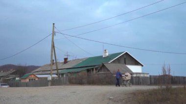 Bir adam bulutlu gökyüzünün altında tozlu bir yolda yürüyor, yeşil çatılı bir binanın önünden geçiyor. Çatıda bir aksesuar var, muhtemelen elektrik ya da rüzgar için bir direk.