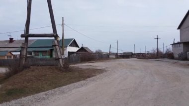 Bulutlu bir gökyüzünün altında evler, elektrik hatları ve telefon direkleriyle kaplı küçük bir köyün içinden geçen toprak yolu olan sakin bir kırsal alan.