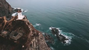 Okyanusun yukarısındaki bir uçuruma tünemiş bir deniz feneri, kıyı ve okyanus manzaralarının akışkan doğal manzarasına tepeden bakan, altında bir plaj ve ufuktaki sıvı denizin ufkuna sahip.