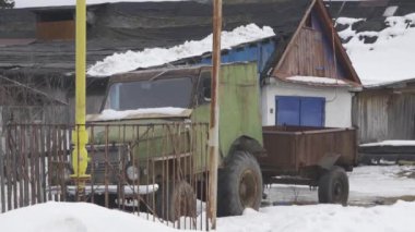 Karavanı olan yeşil bir motorlu araç evin önündeki karda park edilmiş, lastikleri karla kaplı, arka planda bir ağaç ve penceresi var.