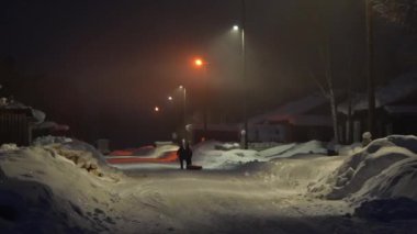 Bir insan gece karlı bir sokakta yürürken, bir sokak lambası sıcak bir ışık saçıyor. Huzurlu karlı ortamda, sakin bir kış akşamının güzelliğini yakalayarak, huzurlu atmosfer güçlenir.