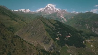 Bir dağ silsilesindeki karlı bir dağın açık bir arkaplan ve bulutlu havadan görüntüsü. Doğal manzara vadiler, tepeler ve otlakları içerir.