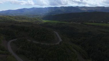 Havadan çekilen bir görüntü, canlı yeşil tepeler ve görkemli dağlar arasındaki dolambaçlı bir yolu yakalar.