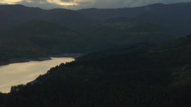 Sakin bir göl yeşil ormanlar ve uzak dağlarla çevrilidir. Hepsi de karamsar atmosferik bir gökyüzünün altında.