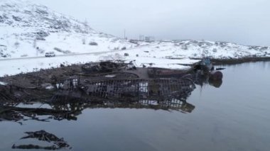 Huzursuz edici bir gemi enkazı, karla kaplı bir arazide güzel, sakin bir su kütlesinin yanında sessizce yatıyor. Arktik Köy Teriberka, Kola Yarımadası, Rusya