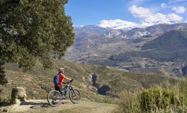 Granada, Anslusia, İspanya yakınlarındaki İspanyol Sierra Nevada 'nın karlı dağlarının altında elektrikli dağ bisikletiyle bisiklet süren hoş, aktif, son sınıf öğrencisi bir kadın.