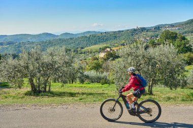 nice senior woman riding her electric mountain bike between olive and cypress trees in the hills near Arezzo in Tuscany,Italy