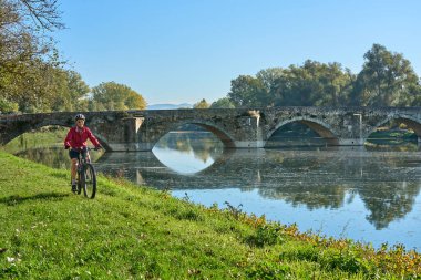 nice senior woman riding her electric mountain at river Arno near Arezzo, with famous bridge of Ponte Buriano in background, Arezzo,Tuscany , Italy