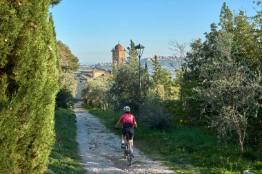 Ghianti bölgesinde zeytin ağaçları arasında elektrikli dağ bisikleti süren hoş yaşlı bir kadın. Arka planda Montepulciano, Toskana, İtalya.
